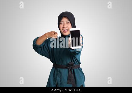 Porträt einer aufgeregten asiatischen muslimischen Frau mit Hijab, die ein Mobiltelefon mit leerem Bildschirm zeigt. Werbemodell-Konzept. Isoliertes Bild auf WHI Stockfoto