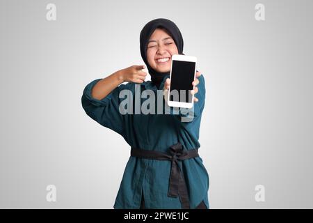 Porträt einer aufgeregten asiatischen muslimischen Frau mit Hijab, die ein Mobiltelefon mit leerem Bildschirm zeigt. Werbemodell-Konzept. Isoliertes Bild auf WHI Stockfoto