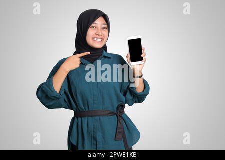 Porträt einer aufgeregten asiatischen muslimischen Frau mit Hijab, die ein Mobiltelefon mit leerem Bildschirm zeigt. Werbemodell-Konzept. Isoliertes Bild auf WHI Stockfoto