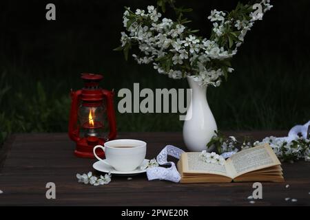 tasse Tee mit Kirschblütendekoration auf dunkelbraunem Holzhintergrund. Schokolade-, Strauß-, Buch- und Kerosinlampe Stockfoto