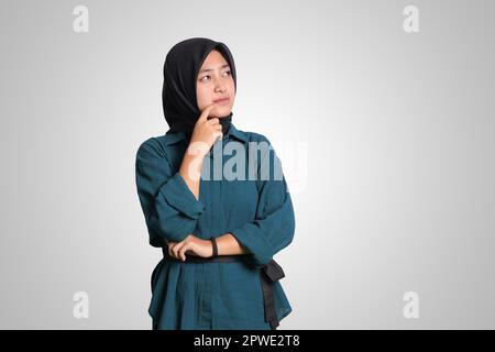 Porträt einer verwirrten asiatischen muslimischen Frau mit Hijab, der vor weißem Hintergrund steht, aufblickt und mit der Hand am Kinn über Fragen nachdenkt Stockfoto