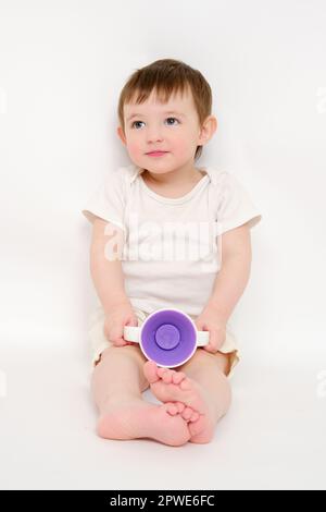 Fröhliches Baby trinkt Wasser aus einer Tasse auf weißem Studiohintergrund. Ruhendes Kind mit Tassensaft in den Händen. Kind etwa zwei Jahre alt (ein Jahr neun Monate) Stockfoto