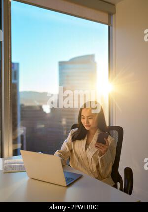 Junge asiatische Geschäftsfrau, die mit einem Telefon-Laptop in einem sonnigen Büro arbeitet, vertikal. Stockfoto