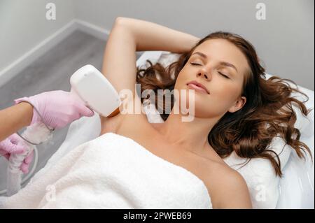 Ästhetische Kosmetologie. Laserepilierung. Eine weiße junge Frau liegt auf einer Couch in einem Schönheitssalon während der Laserhaarentfernung der Achselhöhle Stockfoto