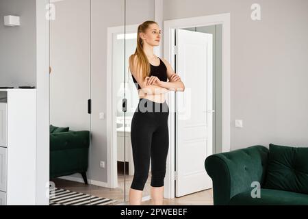 Eine junge Frau in Sportbekleidung steht im Wohnzimmer, die Arme sind auf der Brust gekreuzt Stockfoto