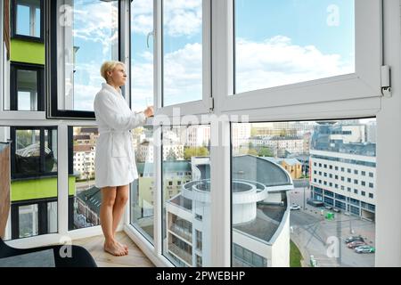 Eine reife, sorgenfreie Frau steht barfuß in einem Bademantel auf ihrem Balkon und genießt einen Orangensaft. Allein in ihrem neuen Hochhaus und mit Blick auf das Haus Stockfoto