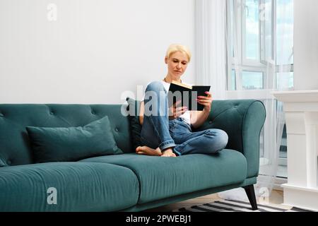 Eine Frau mittleren Alters mit blondem Haar entspannt sich auf dem Sofa in ihrem Wohnzimmer und liest einen Roman. Sie ist lässig gekleidet und genießt ihren Komfort Stockfoto