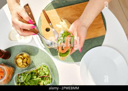 Von oben auf die Hände einer Frau mittleren Alters legt sie salzigen Lachs auf ein Sandwich, während sie Frühstück macht Stockfoto