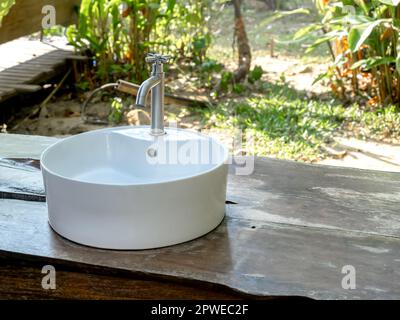 Matt verchromter Wasserhahn auf rundem Keramikbecken Spülbecken auf Holztisch Gartenhintergrund mit Kopierbereich. Modernes Design mit natürlichem Bad Stockfoto