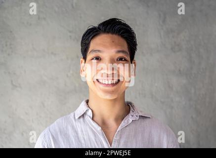 Portrait eines glücklichen Asiaten - selbstbewusster und lächelnder junger asiatischer Mann, der Spaß hat, während er vor der Kamera vor einem grauen Hintergrund posiert - Young Stockfoto