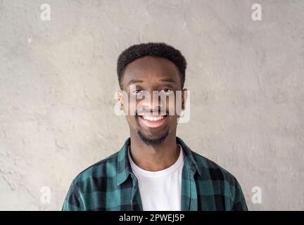 Portrait eines glücklichen afrikaners - selbstbewusster und lächelnder junger schwarzer Mann, der Spaß hat, während er vor der Kamera vor einem grauen Hintergrund posiert - Young Stockfoto