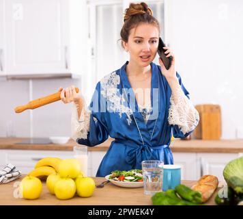 Unzufriedene Frau mit einer rollenden Nadel in den Händen, die in Kitchen telefoniert Stockfoto