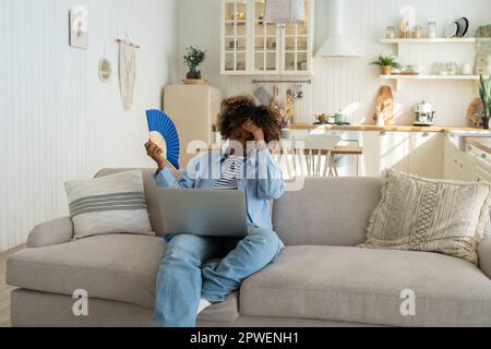 Schwarze Frau leidet unter Hitzschlag, flach ohne Klimaanlage, die den Ventilator winkt, wenn sie zu Hause auf der Couch sitzt Stockfoto