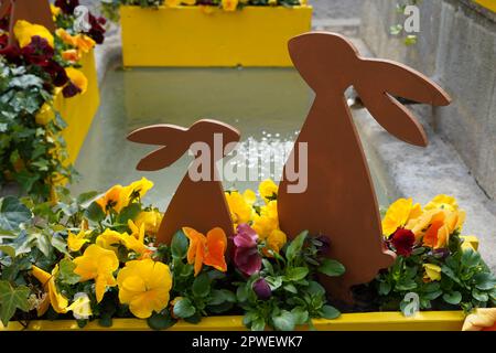 Zwei Osterhasen im Freien, groß und klein, aus Holz und zwischen Blumen platziert. Im Hintergrund befindet sich Springbrunnen. Stockfoto