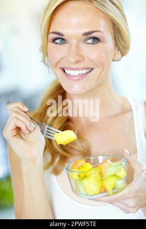 Das ist unglaublich lecker. Attraktive junge blonde Frau, die zu Hause einen köstlichen frischen Obstsalat genießt. Stockfoto