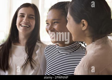 Indische, afrikanische und kaukasische Freundinnen, die lachen, drinnen stehen, dicht beieinander Stockfoto