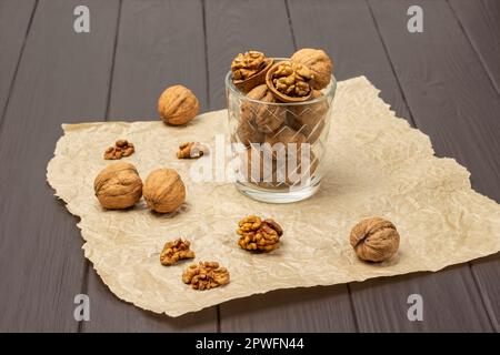 Ganze und gebrochene Walnüsse in einem Glas auf Papier. Walnussschalen und Kerne auf dem Tisch. Dunkler Holzhintergrund. Stockfoto