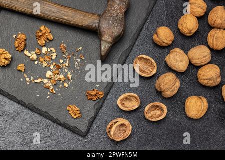 Zerdrückte Walnüsse und ein rostiger, alter Hammer auf einem Steinbrett. Mutternschale und ganze Muttern auf dem Tisch. Flach verlegt. Schwarzer Hintergrund. Stockfoto