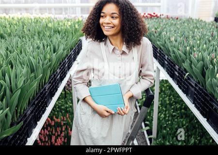 Das junge süße afrikanische Mädchen benutzt ein digitales Tablet, um mit Tulpen im Gewächshaus zu arbeiten. Eine Farmerin mit dunklem Häutchen steht auf einer Trittleiter inmitten von Blumen. Stockfoto