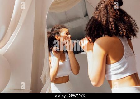 Eine reizende junge Mischrasse in weißer Kleidung steht neben dem Spiegel und reflektiert darin. Süßes, lächelndes schwarzes Mädchen mit lockigem Haar genießt Sonnenlicht in der Nähe des Windos Stockfoto