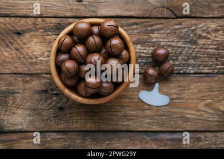 Schüssel mit ganzen leckeren Bio-Macadamianüssen auf Holzhintergrund. Metallschlüssel für Macadamianuss. Macadamia integrifolia in der Schale. Herzhafter Snack. Oben Stockfoto