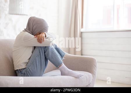 Junge muslimische Studentin im Hijab, sitzt zu Hause auf dem Sofa, ruht sich auf Knien aus und weint. Probleme mit dem Studium, Eltern, Freund. Stockfoto