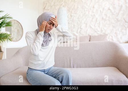 Eine junge Frau in einem Hidschab sitzt zu Hause auf dem Sofa und hält ihren Kopf mit ihren Händen, fühlt starke Schmerzen, gibt sich selbst eine Massage, grimpert von der Spannung. Stress, Sorgen, Depressionen. Stockfoto