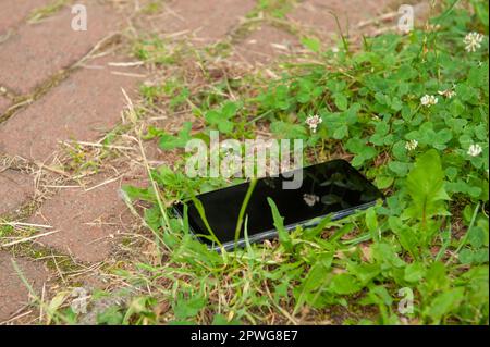 Smartphone liegt auf Gras in der Nähe von Straßenbelägen im Freien. Verloren und gefunden Stockfoto