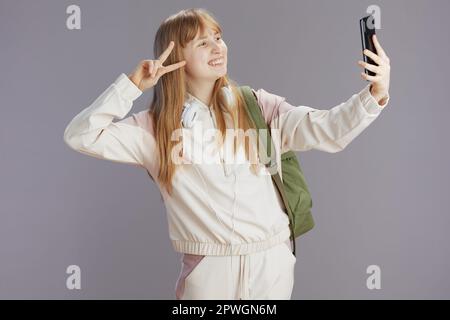 Ein lächelndes, modernes Schulmädchen in beigefarbenem Trainingsanzug mit Rucksack und Kopfhörern, das Selfie macht und das Smartphone gegen Grau verwendet, um Siegesgeste zu zeigen. Stockfoto