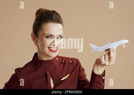 Lächelnde, stilvolle Stewardess isoliert auf beigem Hintergrund mit einem kleinen Flugzeug. Stockfoto