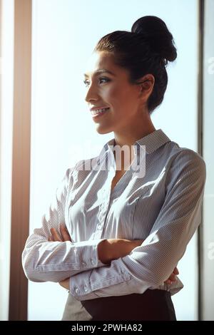 Harte Arbeit hat sie an die Spitze gebracht. Eine attraktive junge Geschäftsfrau, die mit gefalteten Armen im Büro steht. Stockfoto