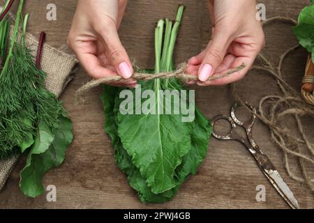 Frau bindet einen Haufen frischer grüner Blätter mit Garn am Holztisch, Draufsicht. Kräuter trocknen Stockfoto