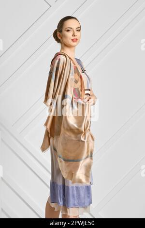Schöne Frau mit Seidenschal auf den Schultern, die im Profil an der weißen Wand steht Stockfoto