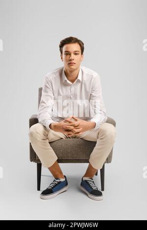 Der nachdenkliche junge gutaussehende Mann in beiger Hose und weißem Hemd sitzt im Studio im Sessel, hat seine Finger verriegelt und schaut geradeaus. Stockfoto