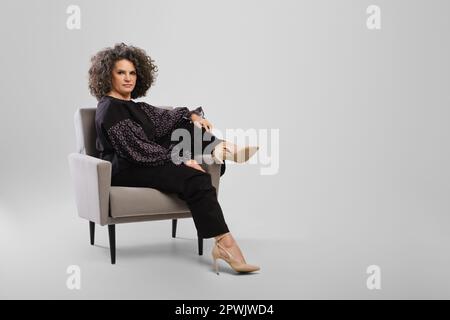 Ältere Frau mit grauem lockigen Haar sitzt in einem Sessel im Studio auf grauem Hintergrund Stockfoto