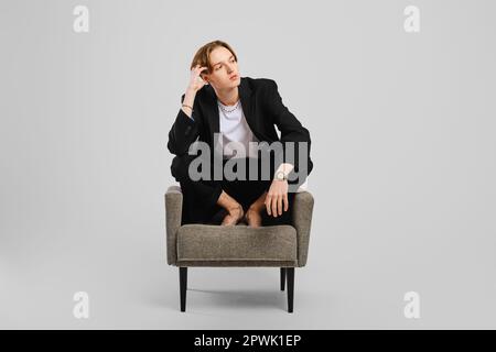 Junger Barfußmann im schwarzen Anzug, der im Sessel im Studio sitzt Stockfoto