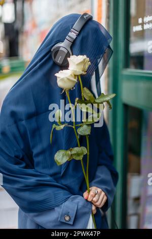 Ein Kopfhörer, der eine muslimische Frau trägt, einen Schleier trägt und Blumen in der Hand hält. Stockfoto