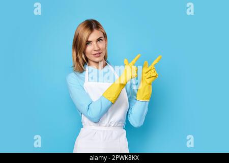 Selbstbewusste, weiße, blonde Frau in gelben Gummihandschuhen, die mit den Fingern auf den Kopierbereich zeigt, isoliert auf blauem Hintergrund. Reinigung Stockfoto