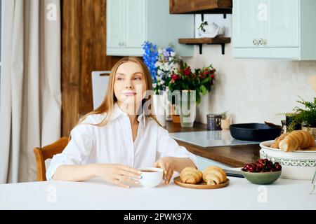 Positive junge Frau sitzt hinter dem Küchentisch mit einer Tasse Kaffee Stockfoto
