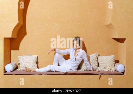 Eine stilvolle Rentnerin mit weißem Hemd und weißer Hose sitzt auf einer orientalischen, wandmontierten Bank Stockfoto