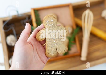 Die Hand einer Frau hält einen Lebkuchen-Mann. Das Kochen von Lebkuchen zu Hause liegt auf einem Holztablett. Urlaubskonzept Stockfoto