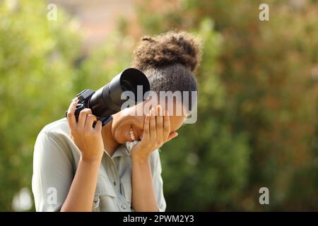 Ein peinlicher schwarzer Fotograf, der lacht und sein Gesicht versteckt, mit einer spiegellosen Kamera in einem Park Stockfoto