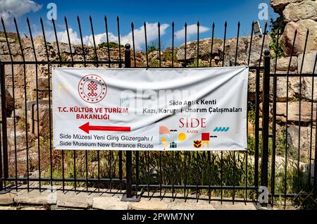 Ein gedrucktes Schild mit Wegbeschreibung zum Side Museum in der türkischen Provinz Antalya (Turkiye) ist an Metallgeländer vor einer zerstörten römischen Mauer gebunden. Die Stockfoto