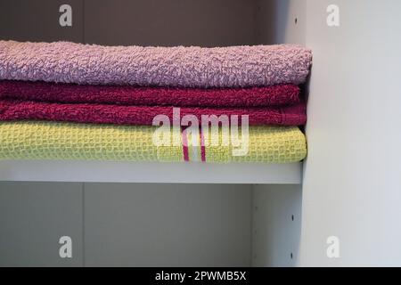 Handtücher auf einem Regal in einem weißen Schrank. Saubere, rosafarbene und gelbe Handtücher, die auf einem Stapel gefaltet sind. Organisation von Haushaltsgegenständen im Badezimmer oder Schrank. Stockfoto