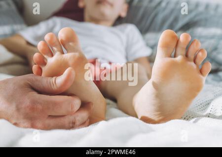 Papa kitzelt die Füße des Kindes. Junge liegt auf dem Bett und spielt mit seinem Vater. Familie, die Spaß zu Hause hat Stockfoto