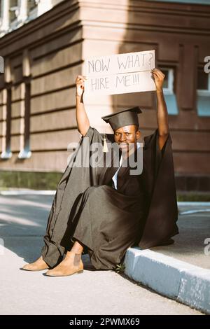 Arbeitsloser Schwarzer, der auf der Straße auf der Suche nach einem Job mit einem hochgezogenen Pappposter sitzt. Universitäts- oder Hochschulstudentin in Graduiertenkleid und Stockfoto