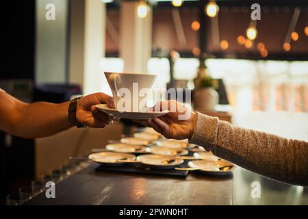 Das ist für Tisch drei hinten. Ein unbekannter Barkeeper, der einem Gast in einem Restaurant Durin eine Tasse Kaffee am Tresen überreicht Stockfoto