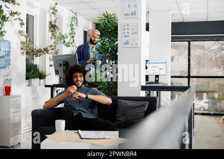 Glücklicher Geschäftsmann, der Geschäftsideen mit der Smartwatch-App aufnimmt, während er im Büro auf dem Sofa sitzt. Lächelnde arabische Führungskraft, die auf einem elektronischen, tragbaren Uhrengerät spricht Stockfoto
