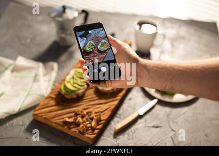 Die Hände fotografieren auf dem Smartphone zwei schöne gesunde Sauerrahm und Avocado Sandwiches liegen an Bord auf dem Tisch. Soziale Medien und Lebensmittel Stockfoto