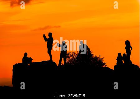 Silhouetten von Menschen auf dem Felsen, die Fotos und Selfies vor dem Hintergrund des orangefarbenen Himmels machen Stockfoto